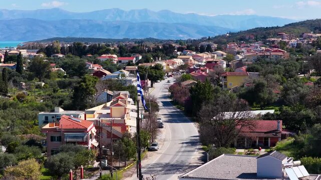 Drone flight over Acharavi town in Corfu, Greece. Beautiful summer view featuring a waving national Greek flag, traditional houses, coastal road, sea, and distant mountain range.