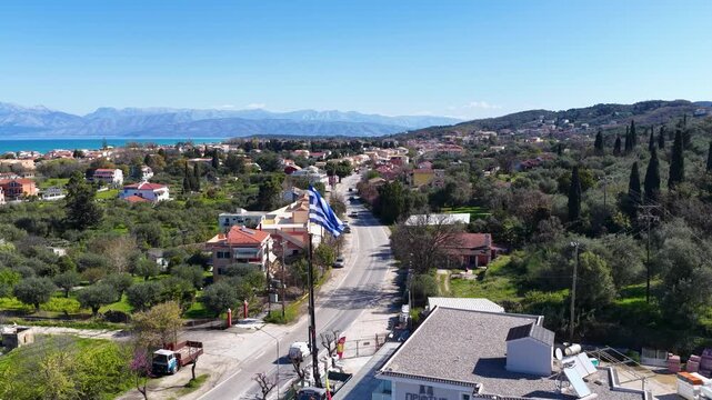 Aerial View of Acharavi North Corfu Greece Wide Shot with Greek Flag Waving Coastal Town Road Traffic Ionian Sea and Mountains