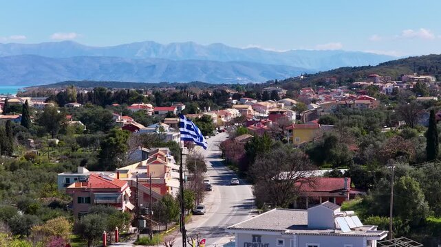 Aerial Drone Flight over Acharavi North Corfu Greece Waving Greek Flag Town Road Traffic Lush Greenery Blue Sea and Distant Mountains
