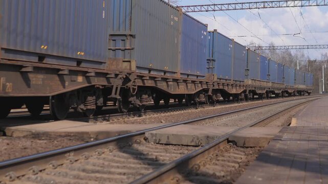 Container Train Rolling Through Rail Yard With Blue Shipping Containers On Flatbed Wagons, Closeup Of Wheels And Tracks, Overhead Catenary Wires, Gravel Ballast And Rusted Metal Details,