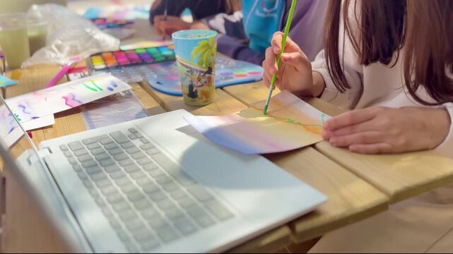 Children painting with watercolor at outdoor table, using laptop for inspiration. Creative learning, art activity, teamwork, education, colorful scene.