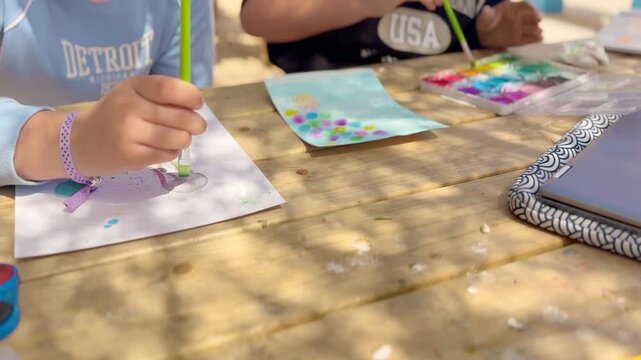 Children painting with watercolor at outdoor table, using laptop for inspiration. Creative learning, art activity, teamwork, education, colorful scene.