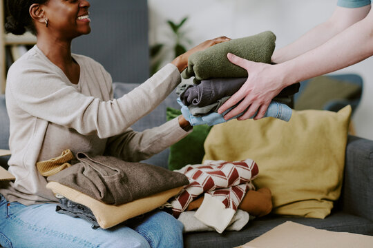 African American young adult woman receiving folded clothes from helper while sorting donation items at home. Supporting charity drive and community aid campaigns, horizontal shot