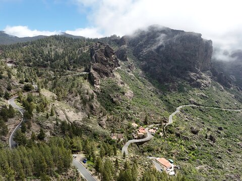 Aerial view of winding roads carve through the lush green landscape, leading to the Roque Nublo peak shrouded in mist, La Culata, Canarias, Spain.