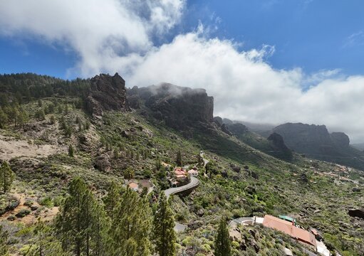 Aerial view of Roque Nublo rising majestically amidst lush green slopes and winding roads under a bright, cloud-strewn sky, La Culata, Canarias, Spain.