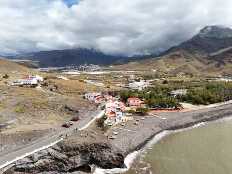 Aerial view of a coastal road winding past white-walled buildings with red-tiled roofs, meeting the dark, rocky shoreline under a sky of swirling clouds, Puerto de la Aldea, Canarias, Spain.