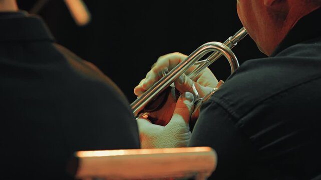 Over the shoulder close-up of a musician playing the trumpet at a live jazz concert. Focus on hands and instrument under warm stage lighting, creating a soulful mood against a dark background