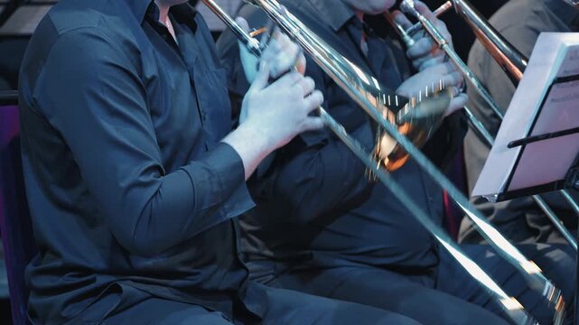 A close-up of musicians in a jazz band brass section playing trombones. The dark, atmospheric scene has cool blue lighting with warm reflections on the golden instruments. Focus on hands sheet music