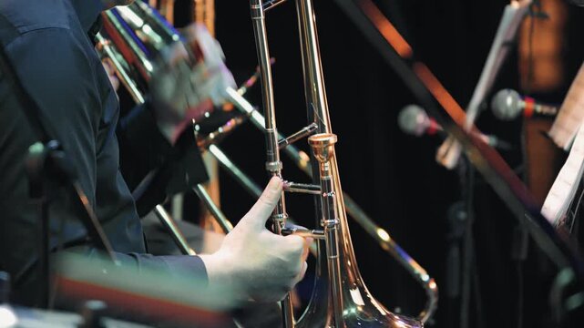 Close-up of a professional trombonist playing in an orchestra. The focus is on the instrument hands, with dynamic stage lighting creating reflections colorful bokeh on a dark background. Moody