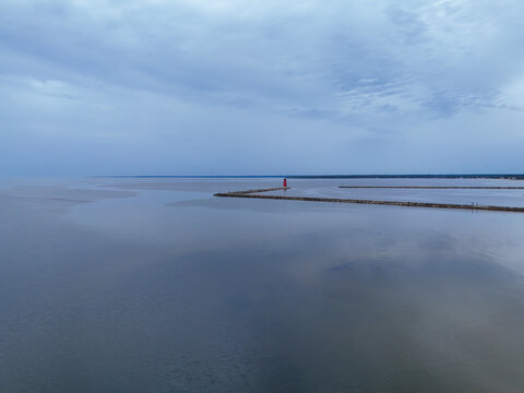 Aerial view of a solitary red lighthouse standing at the end of a long pier, reflecting in the still water under a cloudy sky, Lighthouse, Michigan, United States.