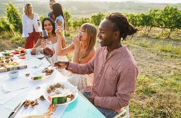 Fototapeta premium Happy adult friends having fun drinking red wine and eating together with vineyard in background - Multiracial people doing party at summer time in countryside resort - Main focus on african man face