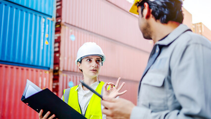Female logistics manager in white hard hat and safety vest holding clipboard talks to male worker...