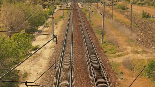 High angle perspective of parallel railroad tracks stretching into the horizon. An empty railway surrounded by dry grass, trees, electric poles under bright daylight. Concept of journey travel