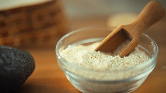 A kitchen countertop features a bowl of sesame seeds beside a spice scoop, accompanied by an unopened green avocado and sliced whole wheat toast bread in the background