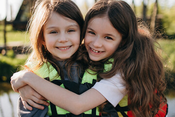 Two happy girls hug each other smiling brightly outdoors near water wearing life jackets © Ananass