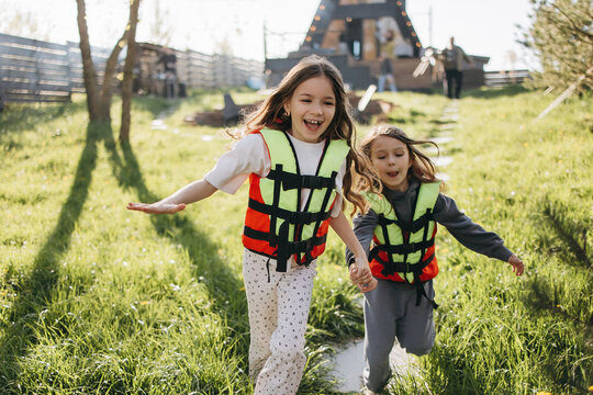Two joyful children run through grass wearing bright life jackets near a wooden cabin on a sunny day