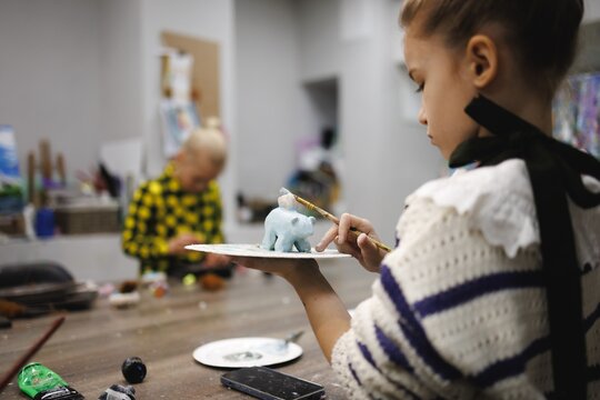 Young girl paints small elephant figurine with brush in art studio while another child works nearby