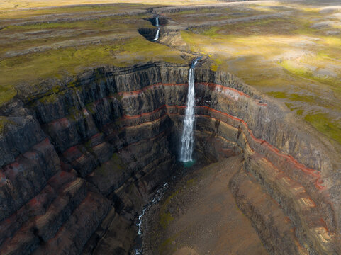 Aerial view of cascading waterfall plunges from layered cliffs into a rugged gorge, contrasting starkly with the flat, grassy plateau above, Egilsstadir, Iceland.