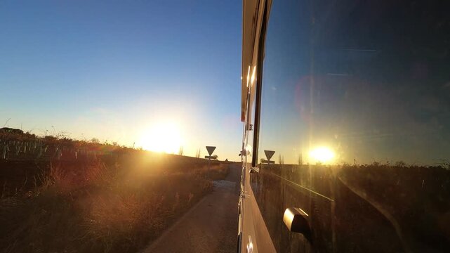 Woman sitting inside camper enjoying travel from passenger seat at sunset, relaxed vanlife lifestyle. Exterior front view of vehicle with road motion, countryside landscape and summer evening light