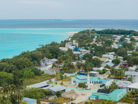 Aerial view of a tranquil island village fringed by turquoise waters, where homes nestle amongst lush greenery under the equatorial sun, Vashafaru Island, Maldives.