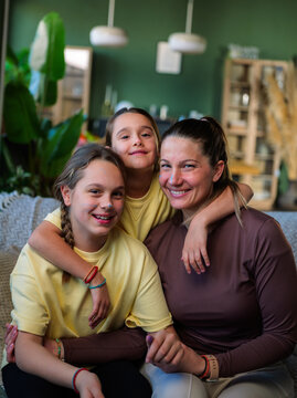 Happy family portrait of a mother embracing her two preteen daughters, all smiling and looking at the camera, relaxing together on a sofa at home, depicting concepts of love and togetherness