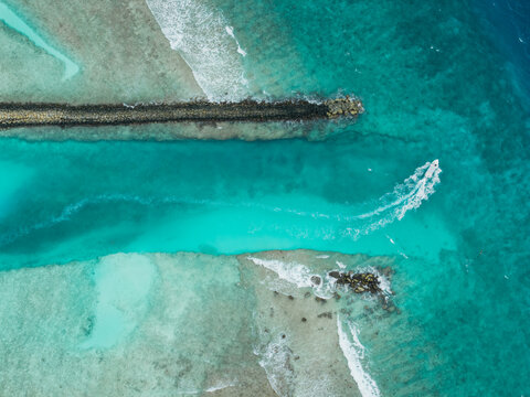 Aerial view of a boat cutting through the turquoise channel bordered by a stone barrier and shallow reefs, Vashafaru Island, Thiladhummathi Atoll, Maldives.