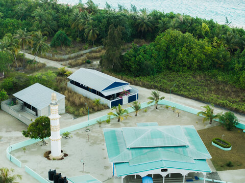 Aerial view of mosque with tall minaret and nearby buildings nestled amid lush greenery, where the land meets the turquoise sea, Vashafaru Island, Maldives.