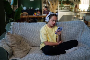 Obraz premium Young girl with cat ear headphones engaging with her mobile phone while sitting comfortably on a sofa at home, enjoying personal entertainment and digital content