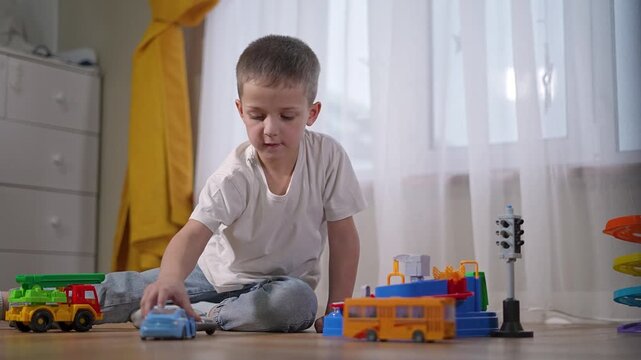 Kid leans floor to grab toy bus and places it care. Son plays calmly beside spiral slide. Learner moves toy truck nearby. Boy drives blue car forward. Boy smiles, learner observes. Kid son build setup