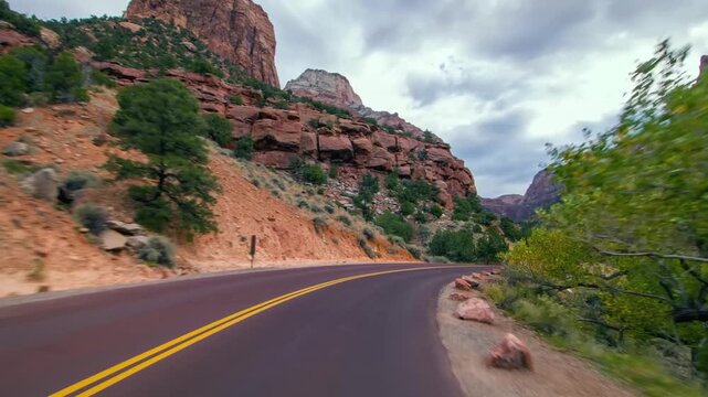 Footage from a car driving along the main road in Zion National Park, Utah. Towering red and orange sandstone cliffs rise on both sides of the winding road, which features yellow double lines. The lan