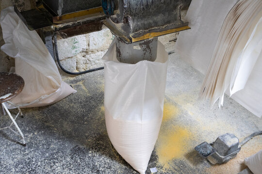 Close-up of an industrial spout filling a white woven sack with yellow cornmeal or flour on a dusty mill floor.