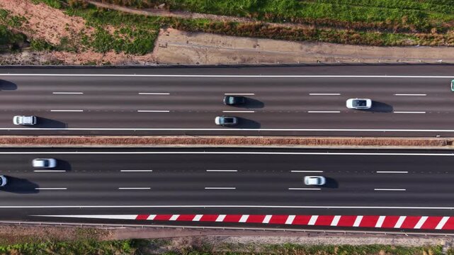 Top-down view of a dual carriageway with cars and trucks moving across white dashed lane markings, red-white barrier chevrons, and a central reservation