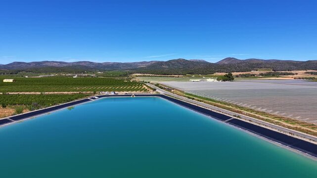 Large turquoise water storage reservoir surrounded by agricultural farmland in rural Spain with mountains and clear blue sky