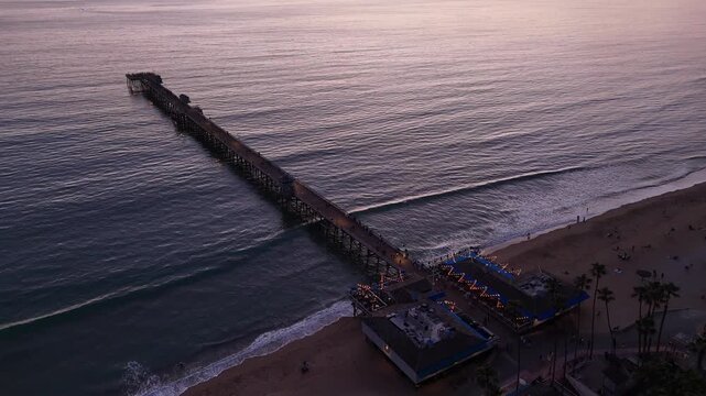 Drone view looking down over San Clemente Pier as gentle waves roll toward the sandy beach at blue hour, with the illuminated pier entrance and calm Pacific water in San Clemente, California.