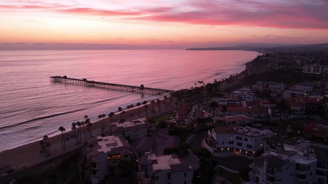 Forward-moving drone shot of San Clemente Pier at sunset with pink clouds, calm Pacific water, palm-lined beach, and coastal homes stretching along the shoreline in San Clemente, California.