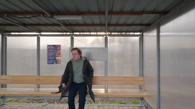 A man on his way somewhere looks at a poster at the bus stop. He sits down on a bench and checks the time