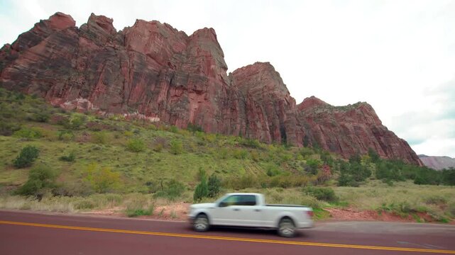 Footage capturing a car driving along the scenic road in Zion National Park, Utah. The shot features towering red sandstone cliffs and lush green vegetation under a cloudy sky