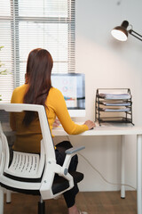 Obraz premium Young woman in a yellow sweater typing at a white desk in a bright home office, focused on remote work and productivity with laptop, documents and daylight streaming through a window