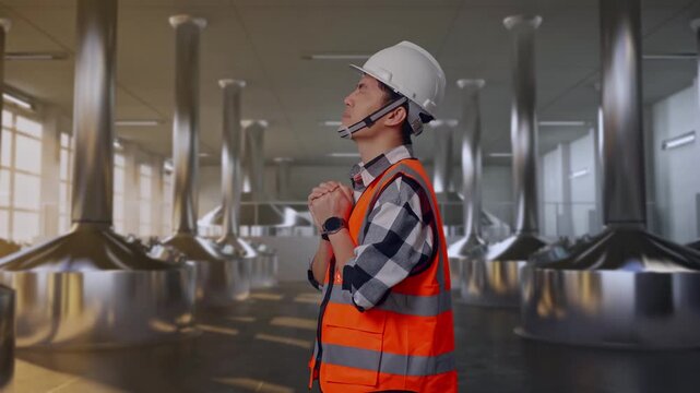 Side View Of Asian Male Engineer With Safety Helmet Pray For Something While Standing at Stainless Steel Brewing Vats in Beer Manufacturing Plant