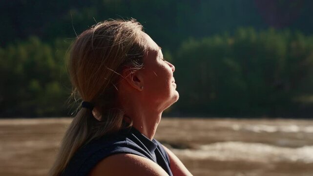 Woman with blonde hair looks upward, eyes closed, smiling peacefully. She stands near a calm river with forested hills behind. Sunlight gently touches her face, creating warm, soft highlights