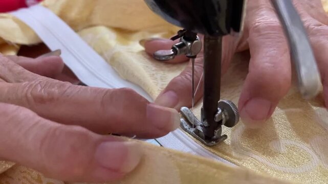 A close-up view of a tailor stitching a white zipper onto yellow patterned fabric using an old manual sewing machine. Focusing on the needlework and garment making.