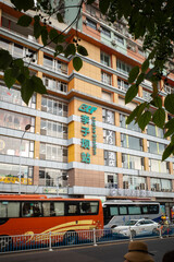 Obraz premium Multi-story building with large green signage. Yellow and gray facade with many windows. Bus and car parked at street level. Green leaves frame the upper left corner. Urban scene with visible traffic