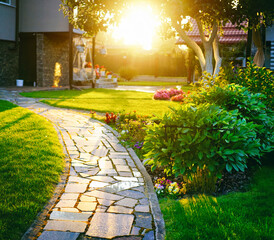 Beautiful stone path leading through a lush green garden at sunset. A peaceful and bright spring nature scene with golden sunlight filtering through the trees.
