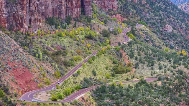A high-angle, panoramic view captures the famous winding switchbacks of the Zion-Mount Carmel Highway as it cuts through the vibrant, rugged landscape of Zion National Park in Utah. The dramatic red r
