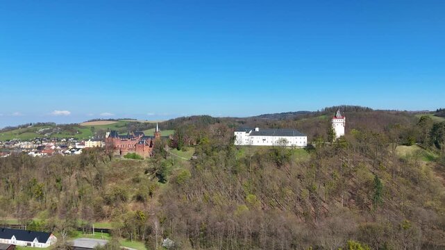 Drone moving sideways and slowly descending captures Hradec nad Moravic&iacute; castle on hill with surrounding countryside, trees and village under clear blue sky.