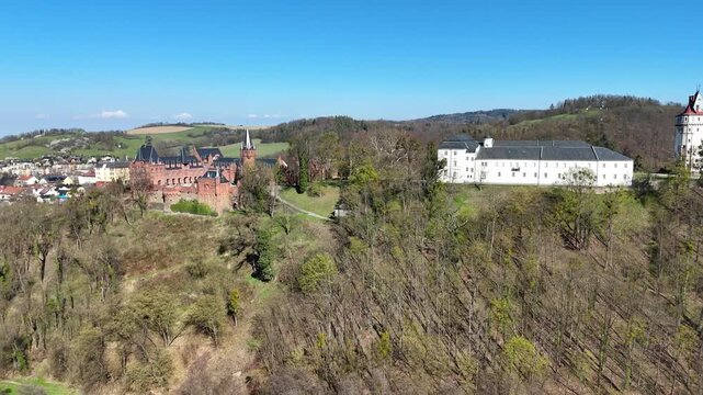 Sideways left movement as drone slowly rotates reveals Hradec nad Moravic&iacute; castle complex with red and white buildings on hill surrounded by trees and scenic countryside.