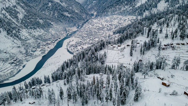 Aerial view of a snow-laden valley with a winding river cutting through the landscape, surrounded by dense pine forests and scattered dwellings, Sharda, Azad Kashmir, Pakistan.