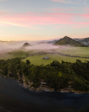Aerial view of the misty, verdant hills and dark sands meeting under a pastel sky, Tongaporutu, Taranaki Region, New Zealand.