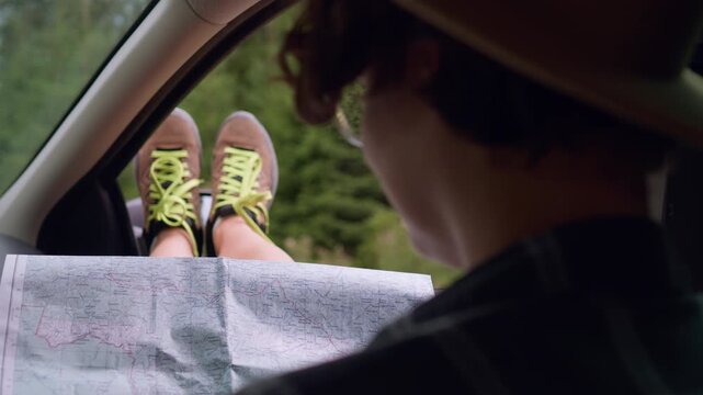 A tourist girl is sitting in a car holding a map looking out for studying a new route for a car trip to the mountains, a traveler sitting with her legs out the car window looking at a map