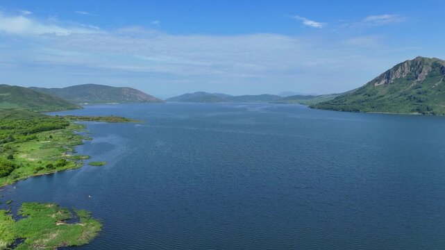 Aerial drone view of the Bukhtarma Reservoir surface with gentle wind ripples. A low mountain covered in lush forest is visible in the distance under the bright summer sun. This footage captures 